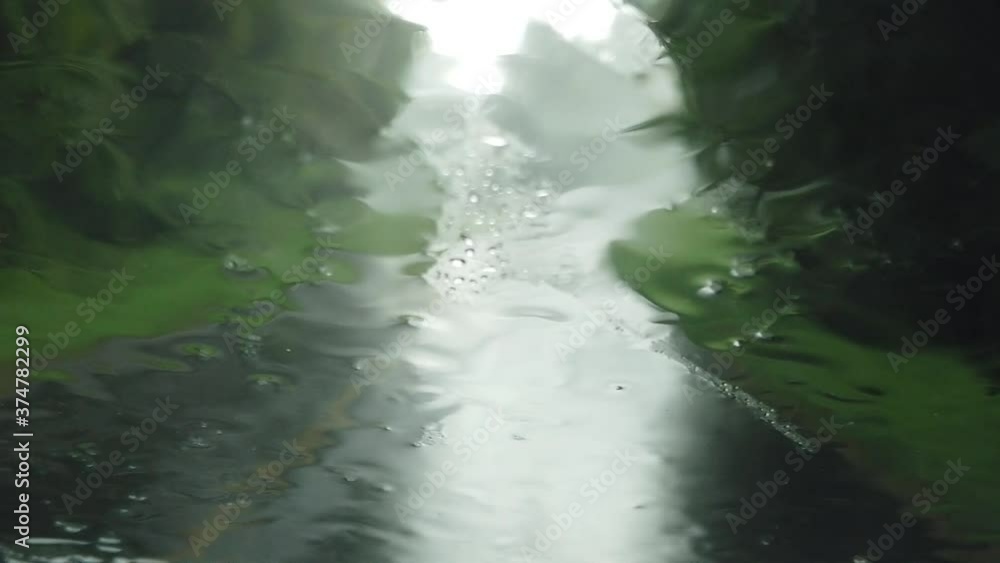 Rain droplets falling, colliding and splashing on car window windshield blurring travel on wet asphalt rural countryside road with green foliage and trees, handheld close up Rain droplets falling, colliding and splashing on car window windshield blurring travel on wet asphalt rural countryside road with green foliage and trees, handheld close up