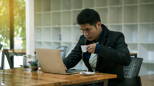 A young Asian businessman who works in front of a laptop and drinks coffee to cure sleepiness in his office.