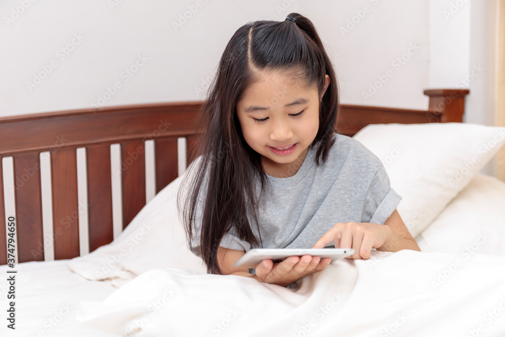 Asian little girl smiling in hands holding and play tablet on bed in bedroom. Little girl enjoy with tablet on bed. Asian girl looking and play tablet. Asian little girl smiling in hands holding and play tablet on bed in bedroom. Little girl enjoy with tablet on bed. Asian girl looking and play tablet.