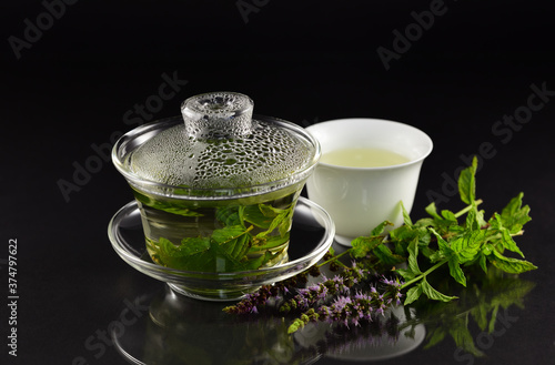 In a Chinese Gaiwan made of glass there is fresh warm peppermint tea in front of a dark background with a teacup, fresh peppermint and flowers
