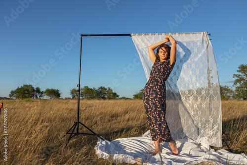 Romantic brunette woman in long dress sitting on old chair in an amazing field, on background white curtains. Warm colors of the sunset.