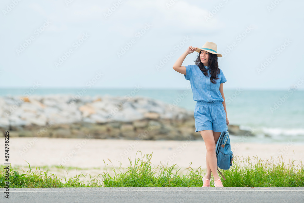 Beach Holiday Happiness. Smiling asian woman in summer casual clothes holding bag and touching her straw hat at the beach enjoying her summer holidays and looking away at copy space.