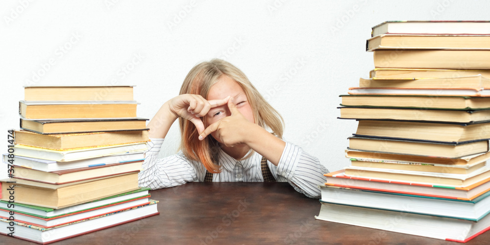 Portrait of a teenage girl sitting at a table tired among the books. Looks through a hand-made frame. Isolated background.