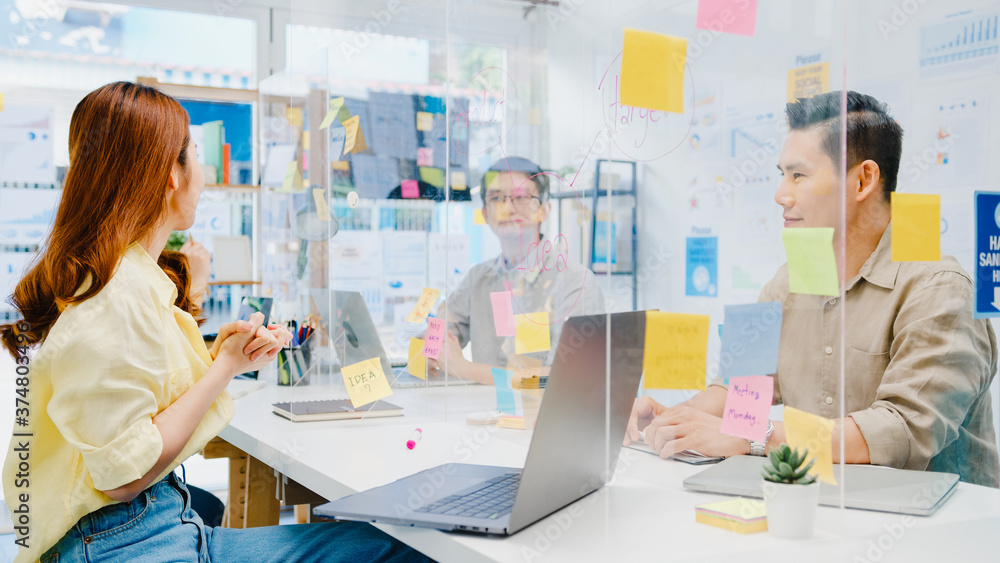 Obraz premium Asia businesspeople discussing business brainstorm meeting together share data and write on acrylic partition stand back in new normal office. Lifestyle social distancing and work after coronavirus.