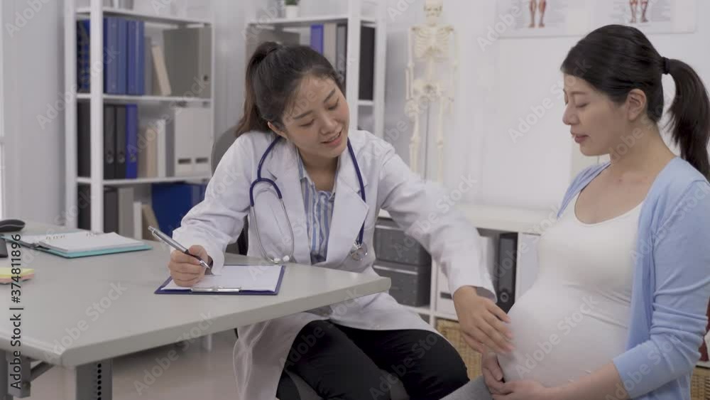 Asian obstetrician sitting leaning toward her patient, touching the swollen belly gently to estimate the fetal size. Asian obstetrician sitting leaning toward her patient, touching the swollen belly gently to estimate the fetal size.