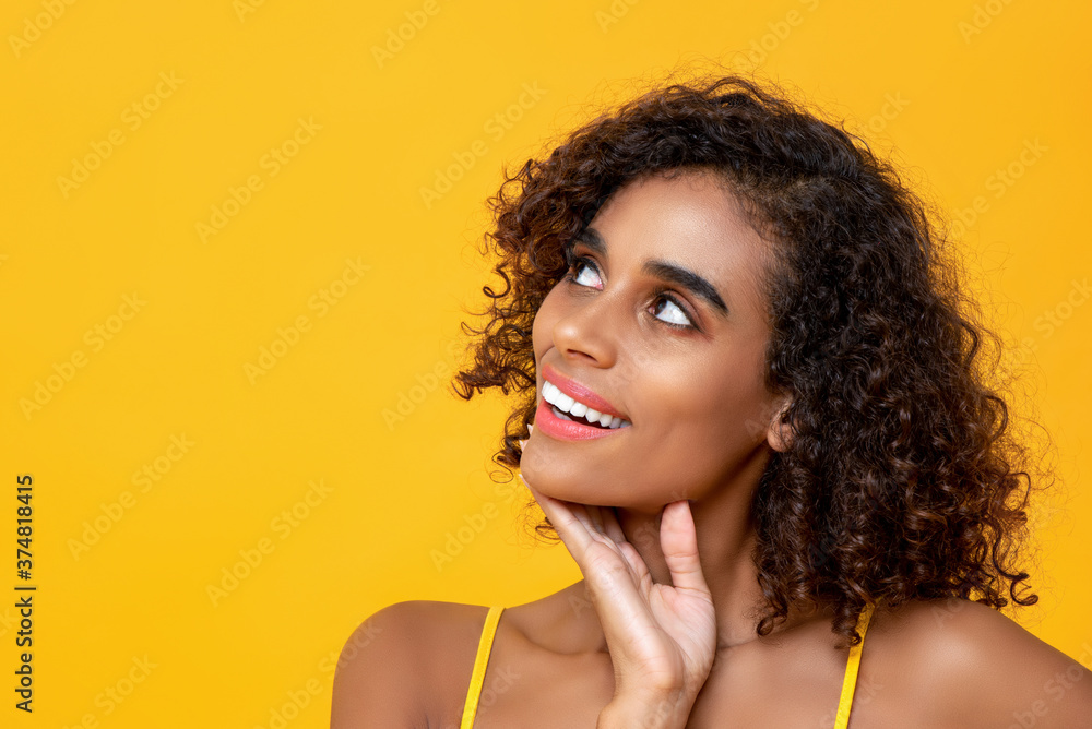 Close up portrait of smiling beautiful Afican American woman thinking looking up isolated on yellow background Close up portrait of smiling beautiful Afican American woman thinking looking up isolated on yellow background