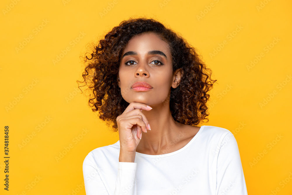 Close up portrait of thinking young African American woman looking at camera with one hand touching chin on studio yellow studio background Close up portrait of thinking young African American woman looking at camera with one hand touching chin on studio yellow studio background