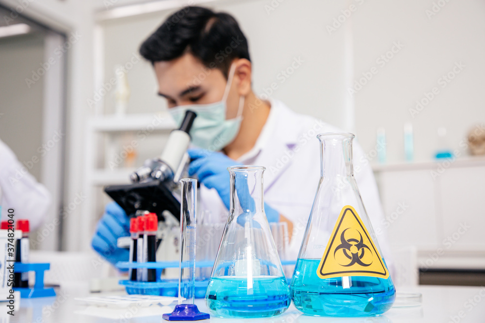 Male Asian Scientist Checking Blood Sample by micro scope. Focus on Tube and Beaker. SARS-CoV-2 , Covid-19 THEME. Male Asian Scientist Checking Blood Sample by micro scope. Focus on Tube and Beaker. SARS-CoV-2 , Covid-19 THEME.