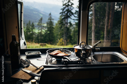 Fotografia Close up on frying pan with organic salmon prepared in inside camper van on campsite in mountains or forest