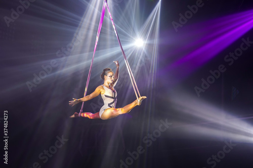 Flexible young woman make performance on aerial straps, flexible split on aerial straps, aerial circus show, purple white light. Flexible woman gymnast on straps, balance split in air