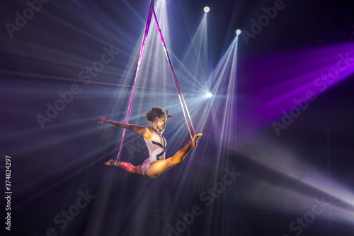 Flexible young woman make performance on aerial straps, flexible split on aerial straps, aerial circus show, purple white light. Flexible woman gymnast on straps, balance split in air