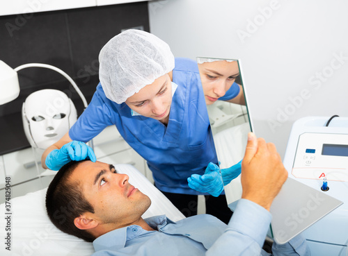 Young man looking at mirror while professional female cosmetician explaining him future hardware facial procedure in cosmetological office.