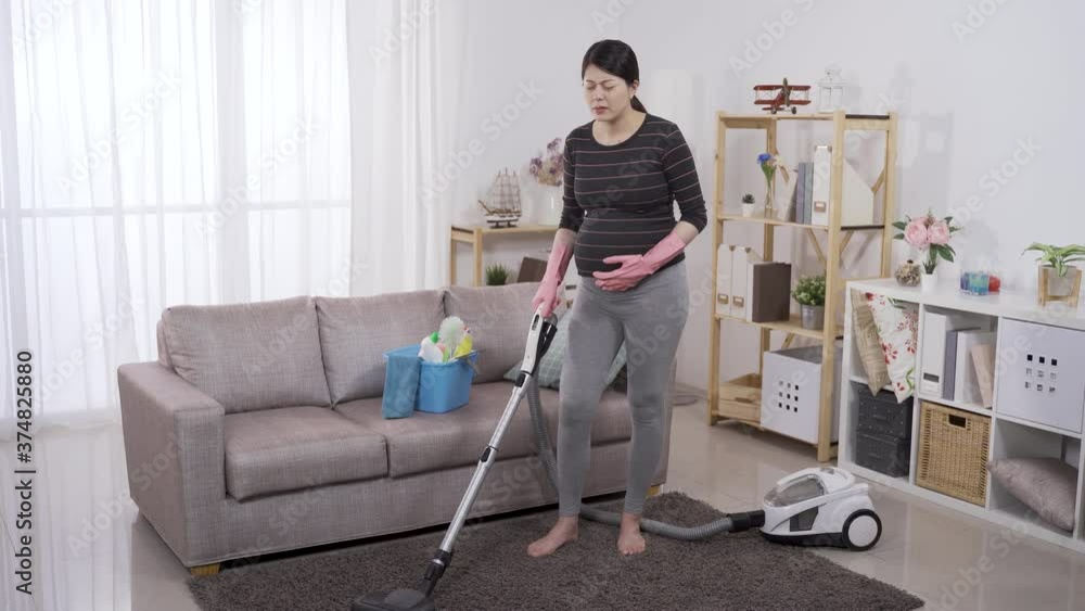 full length asian female in her third trimester feeling sudden painful contraction while cleaning floor with a hoover at home. stroking belly with discomfort facial expression. genuine lifestyle full length asian female in her third trimester feeling sudden painful contraction while cleaning floor with a hoover at home. stroking belly with discomfort facial expression. genuine lifestyle