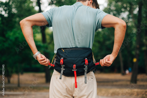 Man in casual clothes stands in the woods during the day and fastens fastex on a waist bag. Tourist with a waist bag on his belt stands in the woods. Back view. photo