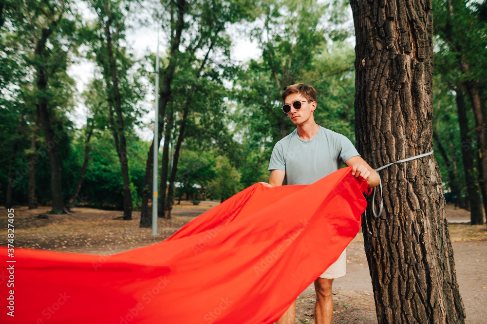 Fototapeta premium Handsome young man touches a red tourist hammock to a tree in the forest with a serious face.