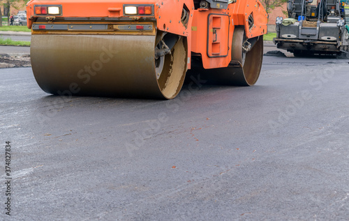 An orange Road roller is moving on freshly paved asphalt. Headlights. Asphalt paver. Glare on the road. Roadway repair concept. Selective focus.