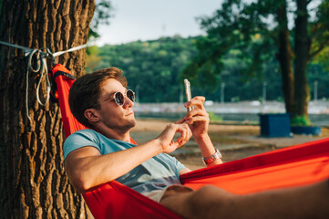 Handsome young man in sunglasses lying at sunset in a hammock and using a smartphone. The guy is resting in a hammock in the park in the evening.