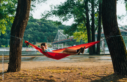 Young man lying in a hammock by the river at sunset and cityscape with a bridge. Relaxing in a hammock on a background of beautiful landscape.