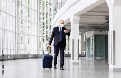 business trip, corporate and pandemic concept - senior businessman wearing face protective medical mask walking with travel bag along office building or airport and checking time