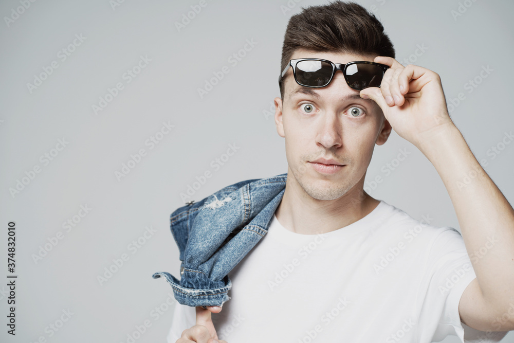 portrait of a surprised young man of Caucasian appearance, holding sunglasses in his hands on his face. the camera is watching. holds on the shoulder of a blue denim jacket portrait of a surprised young man of Caucasian appearance, holding sunglasses in his hands on his face. the camera is watching. holds on the shoulder of a blue denim jacket