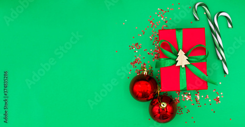 Top view of traditional red christmas present with green bow and little wooden figure of xmas tree  with green glitter or confetti  red balls and candy on green background. Space for text.