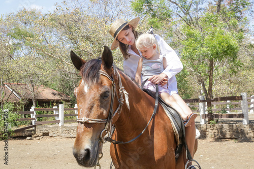 Beautiful young woman riding a horse with her little toddler girl, summer time outdoor activity