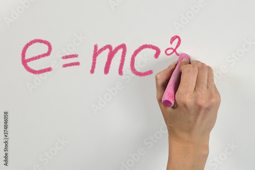 Woman hand with bright pink chalk writing mass-energy equivalence formula on white desk or wall, emc2.