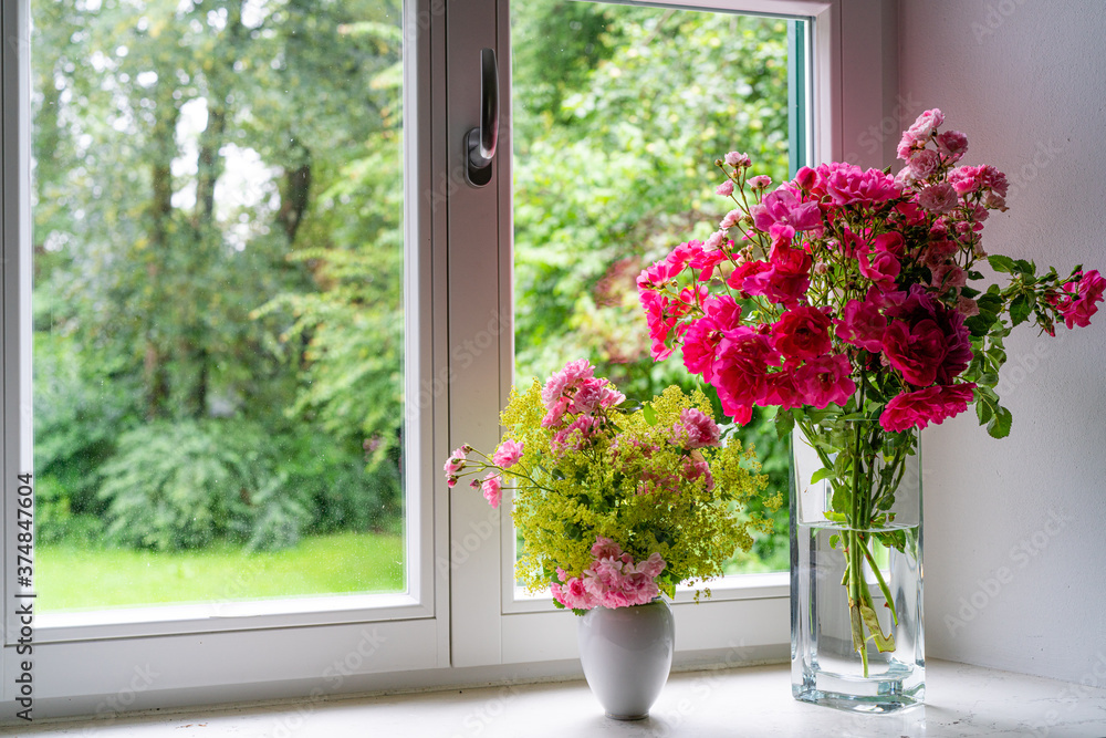 Rosenstrauß mit roten Rosen in Glasvase, auf Fensterbrett mit Blick hinaus in einen weiten Garten