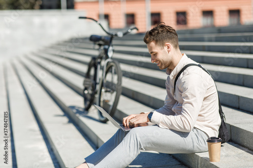 Modern employee sitting on stairs and networking outdoors with takeaway coffee and bike