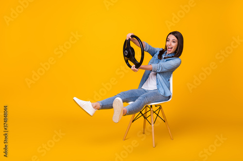 Full body profile photo of pretty funny lady good mood sit chair spread legs playing hold steering wheel riding imagine car wear casual denim shirt shoes isolated yellow color background