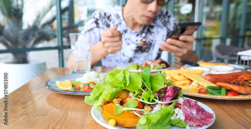 Selective focus of  green salad  plate and  Asian Man using  as looking at his smartphone with having foods of lunch in the restaurant Background