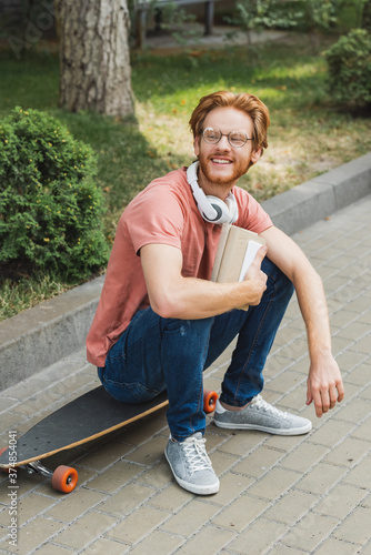 Bearded and redhead student sitting on skateboard and holding book outside