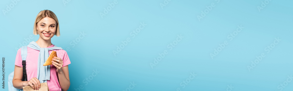 panoramic crop of young blonde student holding paper bag and toast bread on blue panoramic crop of young blonde student holding paper bag and toast bread on blue