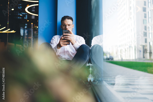 Thoughtful man in eyeglasses sitting near window and using smartphone