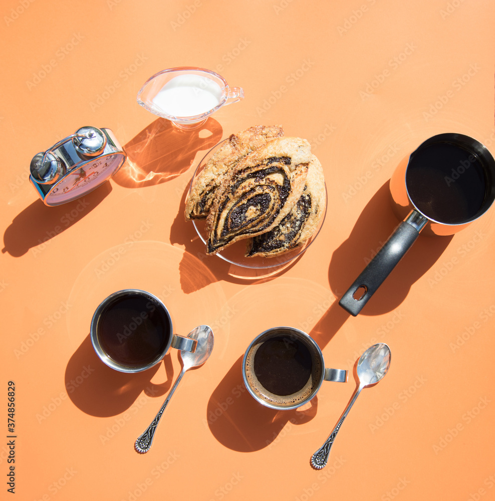 Fototapeta premium On an orange background, there are metal coffee cups, a coffee pot, rolls on a plate, a milkman and an alarm clock, sun glare and shadows from dishes. Horizontal orientation