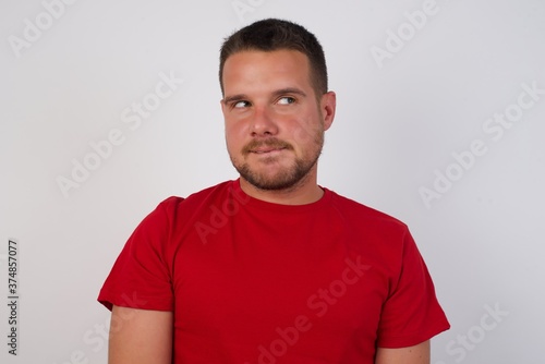 Photo of amazed Young caucasian man wearing red t-shirt over white background bitting lip and looking up to empty space,