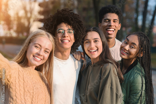 Positive multiracial teen friends taking selfie while walking in park photo
