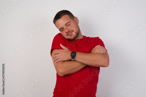 Beautiful young man wearing red shirt over isolated white background Hugging oneself happy and positive, smiling confident. Self love and self care