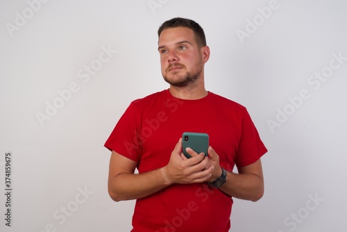 Portrait of Young handsome Cucasian man wearing red shirt standing against white background holding in hands showing new cell