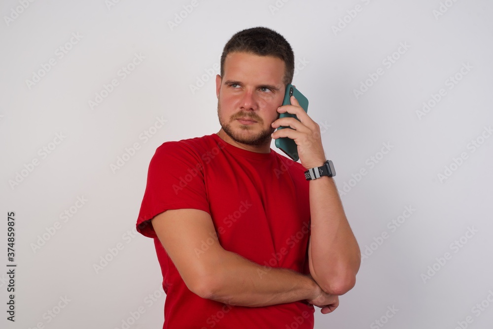 Sad Young handsome Cucasian man wearing red shirt standing against white background talking on smartphone. Communication concept. Sad Young handsome Cucasian man wearing red shirt standing against white background talking on smartphone. Communication concept.