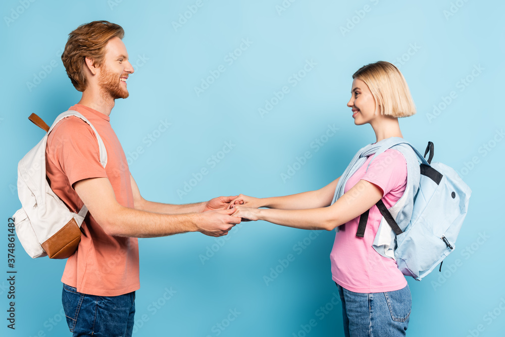 side view of redhead and blonde students with backpacks looking at each other and holding hands on blue side view of redhead and blonde students with backpacks looking at each other and holding hands on blue