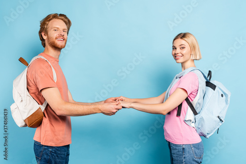 redhead and blonde students with backpacks looking at camera and holding hands on blue