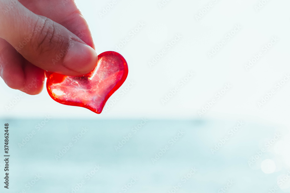 Fototapeta premium A man holds a red glass heart in his fingers against a background of blue sea and blue sky. Beautiful red heart on a Sunny day, the rays of the sun. Bright Sunny sea background.