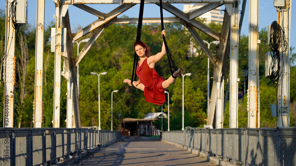 Obraz premium Beautiful sports girl aerialist. circus acrobat on aerial straps, metallic trusses of the bridge. flexible gymnast performs splits against the sky, street show