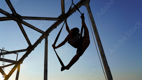Beautiful sports girl aerialist. circus acrobat on aerial straps, metallic trusses of the bridge. flexible gymnast performs splits against the sky, street show