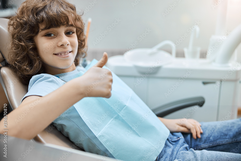 Positive-looking kid holding thumbs up in a dental clinic Positive-looking kid holding thumbs up in a dental clinic