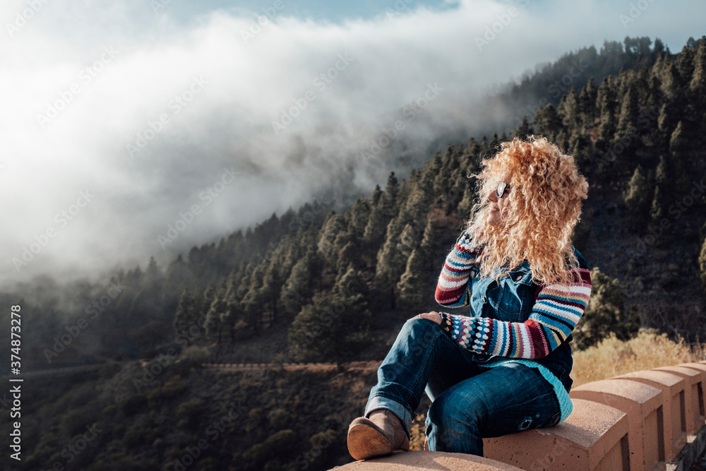 Φωτογραφία Happy beautiful long blonde curly hair woman enjoy and look at the landascape sit down - mountain and forest with clouds in background
