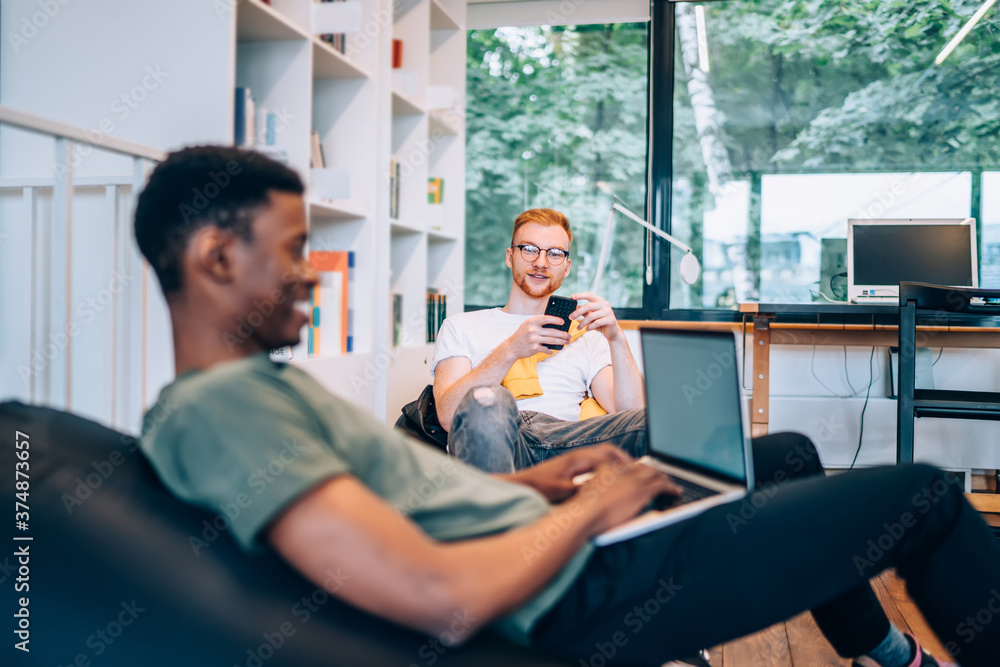 Obraz premium Young man using smartphone near black colleague with laptop