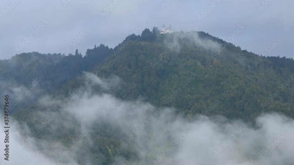 Time lapse low inversion clouds rolling under hill covered with trees. Small catholic church on hilltop Jost in Slovenia. Fog in the valley on a sunny morning. Left truck parallel moving Time lapse low inversion clouds rolling under hill covered with trees. Small catholic church on hilltop Jost in Slovenia. Fog in the valley on a sunny morning. Left truck parallel moving
