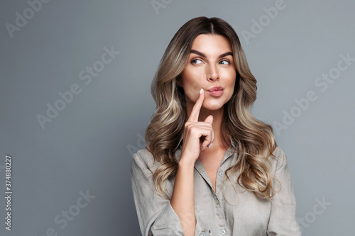 Serious young blonde woman with curly hair dressed in linen shirt thinking, having tricky plan in mind, looking away at blank space for your advertisement, isolated on studio grey background. 